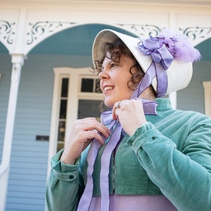 model wearing a turquoise velvet short jacket (Regency Spencer) is standing in front of a blue and white Victorian house and tying the ribbons on her ivory and lavender bonnet