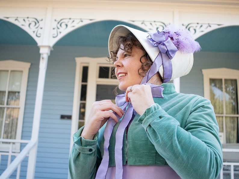 model wearing a turquoise velvet short jacket (Regency Spencer) is standing in front of a blue and white Victorian house and tying the ribbons on her ivory and lavender bonnet