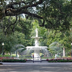 May include: A white fountain with a statue of a woman on top, surrounded by lush green trees and a black wrought iron fence. The fountain is in a park with a paved walkway.