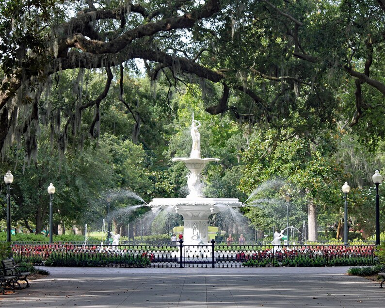 May include: A white fountain with a statue of a woman on top, surrounded by lush green trees and a black wrought iron fence. The fountain is in a park with a paved walkway.