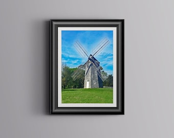 May include: Framed print of a historic windmill on a green field under a blue sky. The windmill is made of weathered wood with large blades. The artwork is in a black frame, suitable for home decor.