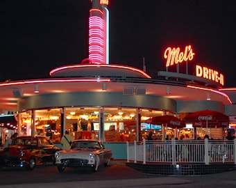 May include: Nighttime exterior shot of Mel's Drive-In, a retro diner with neon signs. The building has a rounded design with a pink and white neon trim. Classic cars are parked in front. The sign reads "Mel's DRIVE-IN".
