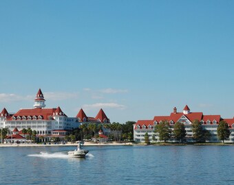 May include: A scenic view of a large white hotel with a red roof and a small boat on the water. The hotel has multiple stories and is surrounded by palm trees and a beach. The sky is clear blue.
