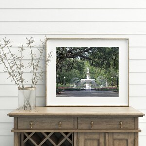 May include: A framed photograph of a white fountain in a park with lush green trees and a white fence. The fountain is surrounded by a grassy area and a paved walkway.