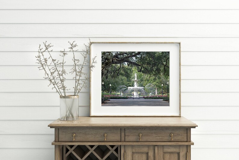 May include: A framed photograph of a white fountain in a park with lush green trees and a white fence. The fountain is surrounded by a grassy area and a paved walkway.