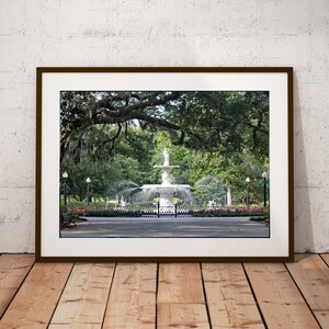May include: A framed photograph of a white fountain in a park with lush green trees and a brick walkway. The fountain is surrounded by a black metal fence and has a statue of a woman on top.