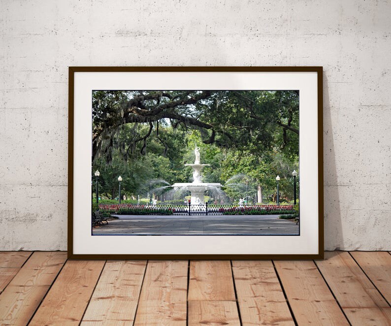 May include: A framed photograph of a white fountain in a park with lush green trees and a brick walkway. The fountain is surrounded by a black metal fence and has a statue of a woman on top.