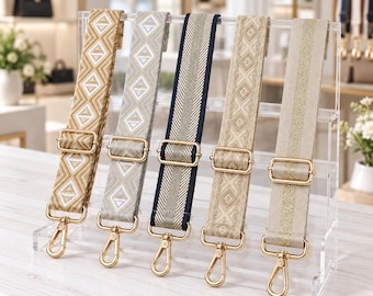 May include: Five adjustable bag straps displayed on a clear acrylic stand. Each strap features gold-tone metal hardware, including a sliding buckle and a lobster clasp. The straps vary in colour and pattern, including geometric designs and metallic stripes.