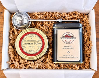 May include: A gift box containing a silver metal tea infuser, a round gold tin labeled Crumpets & Jam 100% soy wax net wt 2 oz / 57 g, and a black rectangular tin labeled The Underground Grocer Earl Grey Premium Black Tea net wt 2 oz / 57 g.