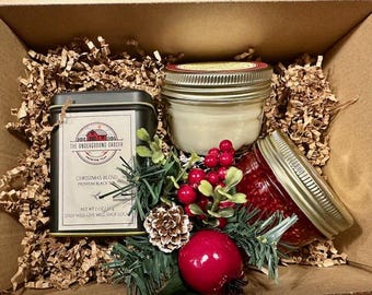 May include: A gift box containing a black tin of The Underground Grocer Christmas Blend tea, a glass jar with a white candle, and a glass jar of red jam. The items are nestled in brown paper filler with a decorative pine sprig, red berries, and a pinecone.