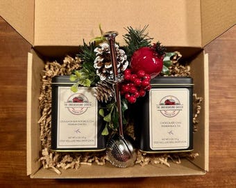 May include: A gift box containing two black rectangular tea tins labeled The Underground Grocer, one with Cinnamon Bun Rooibos Chai and one with Chocolate Chai, each 2 oz/57 g. A silver metal tea infuser sits between them, topped with a pinecone and red berries.