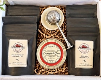 May include: A gift box containing four black pouches of The Underground Grocer tea, including English Breakfast and Earl Grey, a round Crumpets & Jam soy candle, and a silver metal tea infuser with a small white ceramic saucer.