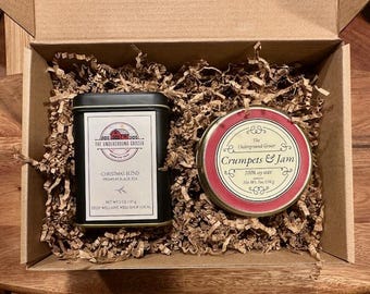 May include: A cardboard gift box containing a rectangular black tin of Christmas Blend premium black tea, net weight 2 oz / 57 g, and a round red tin of Crumpets & Jam 100% soy wax candle, net weight 7 oz / 198 g, nestled in brown paper shred.