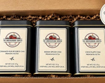May include: A cardboard gift box containing three black rectangular tea tins labeled The Underground Grocer. The labels feature a red barn logo and the text Cinnamon Run Rooibos Chai, Indian Spiced Chai, and Chocolate Chai, each net 2 oz.