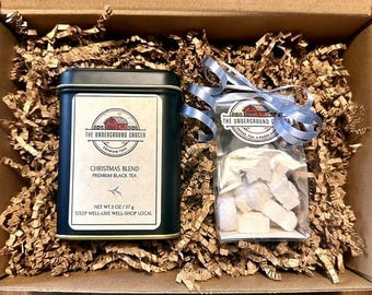 May include: A dark green rectangular tea tin labeled The Underground Grocer Christmas Blend Premium Black Tea Net Wt 2 oz / 57 g and a clear plastic bag of white star-shaped sugar pieces tied with a light blue ribbon inside a cardboard box.