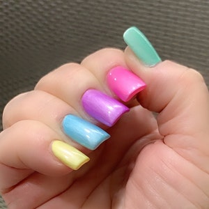 May include: A hand with five fingernails painted in a rainbow gradient. The nails are painted in shades of yellow, light blue, purple, pink, and light green.