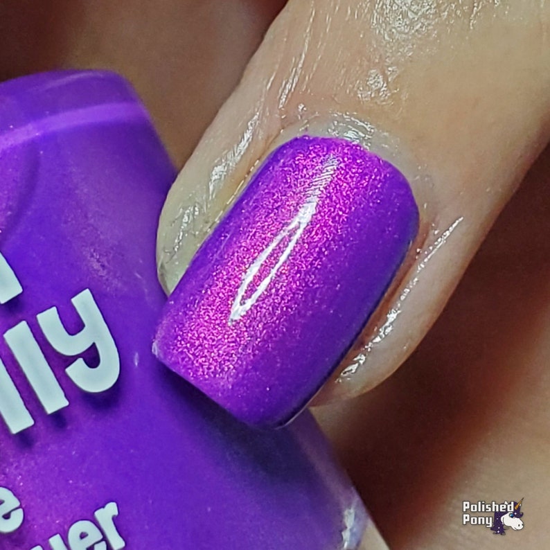 Puede incluir: Primer plano de una u&ntilde;a pintada con un vibrante esmalte de u&ntilde;as morado. El esmalte tiene un efecto brillante y con purpurina. Una botella de esmalte de u&ntilde;as morado a juego es parcialmente visible en el fondo. La marca "Polished Pony" es visible.