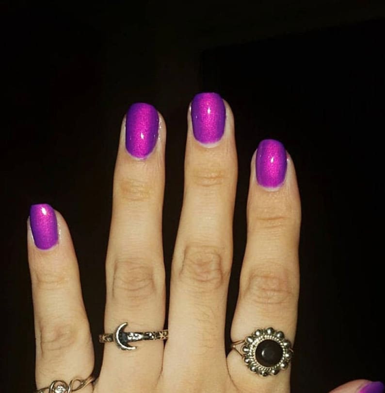 Puede incluir: Primer plano de una mano con cinco dedos, cada u&ntilde;a pintada con un esmalte de u&ntilde;as morado brillante. La mano est&aacute; adornada con varios anillos, incluyendo uno con forma de media luna y otro con dise&ntilde;o floral.