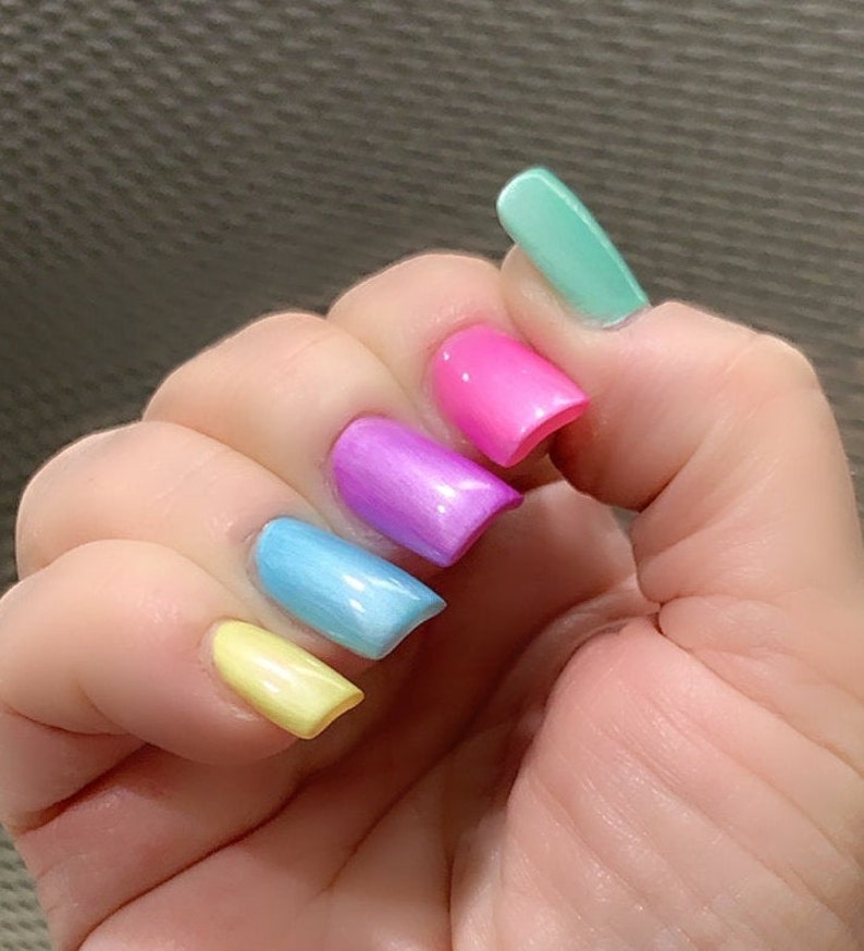 May include: A hand with five fingernails painted in different pastel colors: yellow, light blue, purple, pink, and light green.