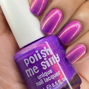 Puede incluir: Un frasco de esmalte de u&ntilde;as &uacute;nico "Polish Me Silly" en un tono p&uacute;rpura vibrante, sostenido en una mano con u&ntilde;as pintadas en un color a juego. Las u&ntilde;as muestran un efecto degradado, que va del rosa al p&uacute;rpura. El frasco tiene una tapa blanca.