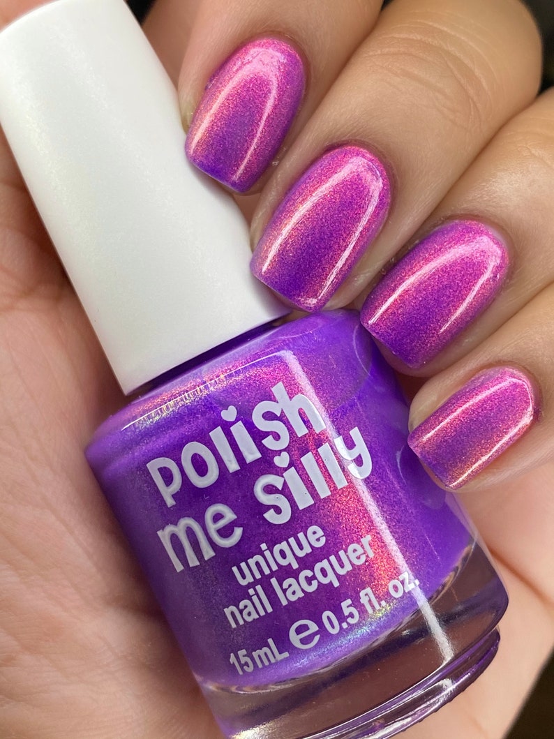 Puede incluir: Un frasco de esmalte de u&ntilde;as &uacute;nico "Polish Me Silly" en un tono p&uacute;rpura vibrante, sostenido en una mano con u&ntilde;as pintadas en un color a juego. Las u&ntilde;as muestran un efecto degradado, que va del rosa al p&uacute;rpura. El frasco tiene una tapa blanca.