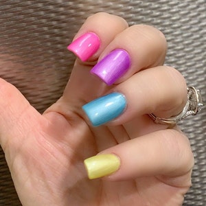 May include: Close-up of a hand with four fingernails painted in different colors: pink, purple, blue, and yellow.