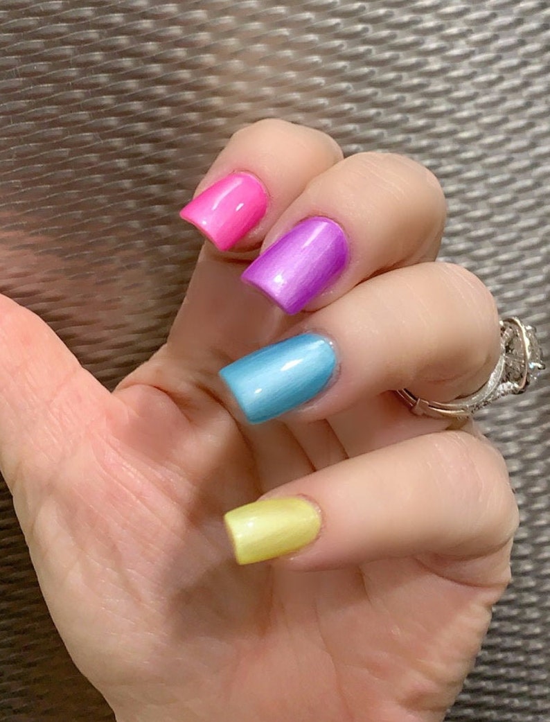May include: Close-up of a hand with four fingernails painted in different colors: pink, purple, blue, and yellow.