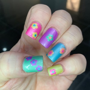 May include: Close-up of a hand with five fingernails painted in different colors with flower designs. The nails are painted in pink, purple, blue, green, and yellow. Each nail has a different flower design with yellow, pink, and blue flowers.