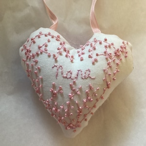 May include: A white heart-shaped pillow with pink embroidery. The pillow has the word "Nana" embroidered in the center.