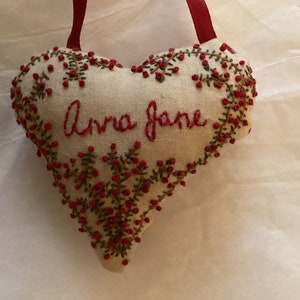 Op de afbeelding: Hartvormig, crèmekleurig stoffen ornament met rode geborduurde tekst "Anna Jane". Het hart is afgezet met groene ranken en rode bessen en heeft een rood lint om op te hangen.