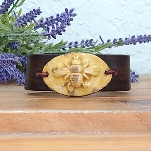 May include: A brown leather bracelet with a yellow ceramic bee charm. The bee is raised and has a detailed design.
