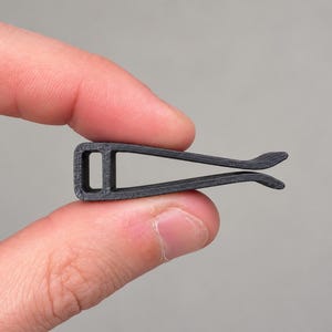 May include: A small, black, 3D-printed plastic clip featuring a rectangular loop at one end and two slightly curved, flexible arms that taper toward the open end. The object has a matte, textured surface finish consistent with additive manufacturing.