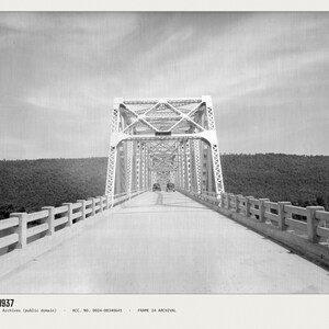 May include: A black and white archival photograph of a steel truss bridge from 1937. The image shows a long, straight road leading into the bridge structure, flanked by concrete railings. Two vintage cars are visible in the distance on the road.