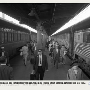 Peut inclure: Une photographie en noir et blanc montrant des passagers et des employ&eacute;s de train marchant sur un quai &agrave; Union Station, Washington, D.C. en 1963. Des personnes portent des bagages entre deux grands trains gar&eacute;s sous un toit de gare couvert.