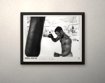 May include: A black and white framed photograph of a boxer training with a heavy bag. The text at the bottom reads CAMEROON - BOXING TEAM, along with additional small print. The image is enclosed in a simple black frame with a white mat border.