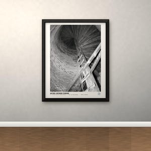 May include: A black and white framed print titled HISTORIC LIGHTHOUSE STAIRCASE, featuring a low-angle view of a spiraling wooden staircase inside a stone tower. The composition captures the vertical ascent of the stairs against the textured wall.