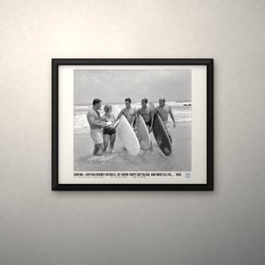 May include: A black and white framed photograph depicting four individuals with surfboards on a beach. The text at the bottom reads: SURFING—CAPTAIN RODNEY BOTHELO, 1ST SHORE PARTY BATTALION, AND MISS ELLI VA... 1966. The print is in a black frame.