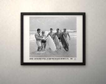May include: A black and white framed photograph depicting four individuals with surfboards on a beach. The text at the bottom reads: SURFING—CAPTAIN RODNEY BOTHELO, 1ST SHORE PARTY BATTALION, AND MISS ELLI VA... 1966. The print is in a black frame.