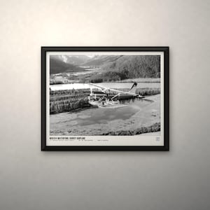 Peut inclure: Une impression encadr&eacute;e en noir et blanc montrant un hydravion survolant un paysage de montagnes, d'arbres et d'eau. Le texte en bas indique WDE154 WATERFOWL SURVEY AIRPLANE, U.S. FISH AND WILDLIFE SERVICE, DEC. 10, 1953, ALASKA, PHOTO BY ARCHIVE.