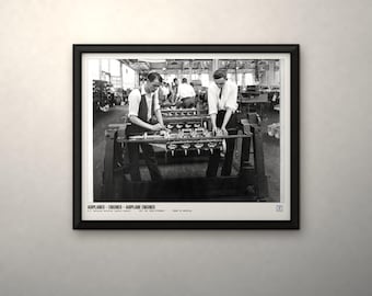 May include: A black and white framed photograph depicting two workers assembling an airplane engine in a factory setting. The print includes the text AIRPLANES - ENGINES - AIRPLANE ENGINES at the bottom, capturing a historical industrial scene.