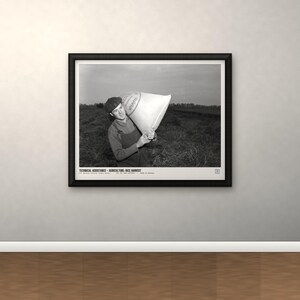 May include: A black and white framed photograph depicting a person in overalls and a cap carrying a large sack in a field. The print includes the text: TECHNICAL ASSISTANCE - AGRICULTURE: RICE HARVEST. The frame is black with a white mat border.