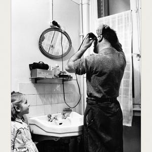 May include: A black and white photograph showing a person using an electric hair clipper in front of a bathroom mirror while a child watches. A shelf holds a box labeled PHILIPE, and the bottom text reads SOLLAC STEEL MILL.