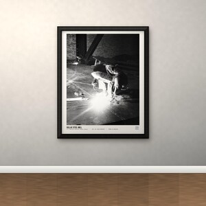 May include: A black and white framed print titled DOLLAC STEEL MILL, featuring a high-contrast photograph of a person welding in an industrial setting with bright sparks, set within a simple black frame on a neutral wall.