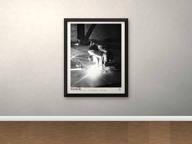 May include: A black and white framed print titled DOLLAC STEEL MILL, featuring a high-contrast photograph of a person welding in an industrial setting with bright sparks, set within a simple black frame on a neutral wall.