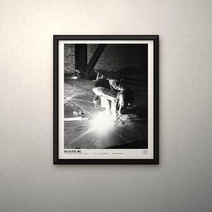 May include: A black and white framed print titled SOLLAC STEEL MILL, featuring a worker welding in a dark industrial space. The bright light from the welding torch illuminates the floor, while the text at the bottom provides details about the scene.