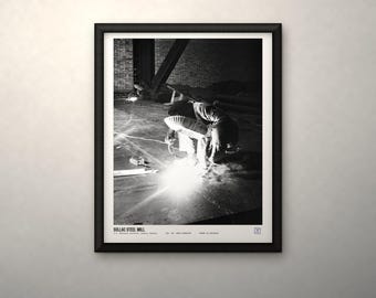 May include: A black and white framed print titled SOLLAC STEEL MILL, featuring a worker welding in a dark industrial space. The bright light from the welding torch illuminates the floor, while the text at the bottom provides details about the scene.