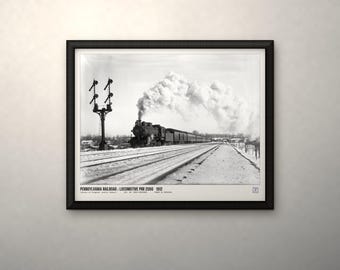 May include: A black and white framed print featuring a steam locomotive on snowy tracks with a signal post. The text at the bottom reads: PENNSYLVANIA RAILROAD : LOCOMOTIVE PRR 2086 1912. The image captures a train emitting smoke in a winter landscape.