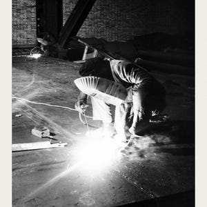 May include: A black and white photograph of a worker wearing a welding helmet and protective clothing, kneeling while welding a large metal surface. The image includes the text SOLLAC STEEL MILL, U.S. National Archives, and ACC. NO. 0024-19982399.