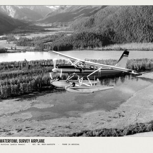 Peut inclure: Une photographie en noir et blanc d'un hydravion survolant une rivi&egrave;re et une for&ecirc;t. Le texte en bas indique WOE154 WATERFOWL SURVEY AIRPLANE, U.S. National Archives (public domain), ACC. NO. 0024-66693378, FRAME 24 ARCHIVAL.