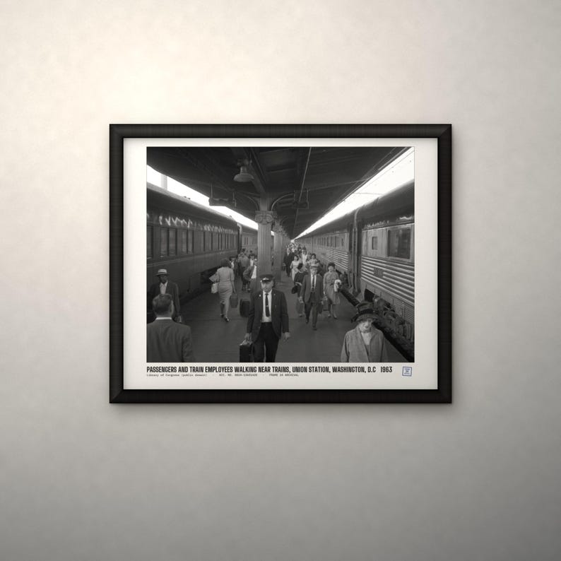 Peut inclure: Une photographie encadr&eacute;e en noir et blanc de la gare Union Station &agrave; Washington, D.C. en 1963, montrant des passagers et des employ&eacute;s marchant entre les trains. Le texte en bas indique : PASSENGERS AND TRAIN EMPLOYEES WALKING NEAR TRAINS, UNION STATION, WASHINGTON, D.C. 1963.